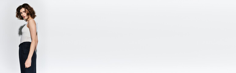 A pretty young woman in black pants and white tank top striking a pose in front of a white wall in a studio setting.