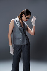 A stylish young woman is posing elegantly in a gray suit with a vest and white gloves in a studio setting against a gray background.