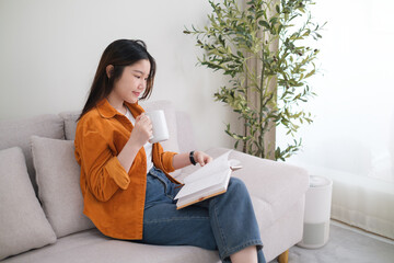 Happy young woman wearing casual clothes drinking coffee and reading book on grey sofa at home.