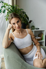 A young woman in activewear sits on a chair in a modern apartment, practicing yoga.