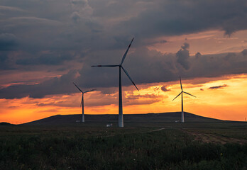 Obraz premium Multiple wind turbines silhouetted against a vibrant sunset sky. The turbines stand tall against the backdrop of orange and red clouds.