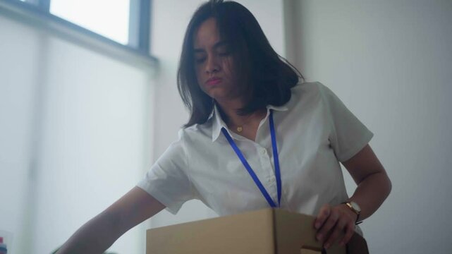 Asian woman packing her belongings into a cardboard box, indicating her resignation from work. She looks focused as she organizes her personal items, including books, in an office setting