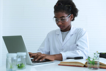 Young curly-haired black girl using laptop to find scientific information about green plants in laboratory.