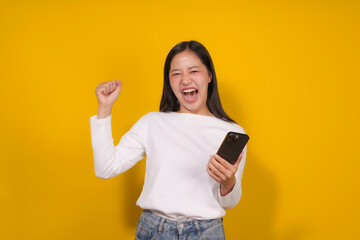 Excited young woman is raising her fist in the air while holding her smartphone, expressing joy and excitement on a vibrant yellow background