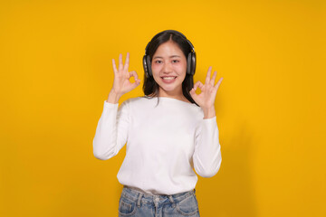 Young woman is enjoying music on her headphones while giving the ok sign with a smile, isolated on a vibrant yellow background