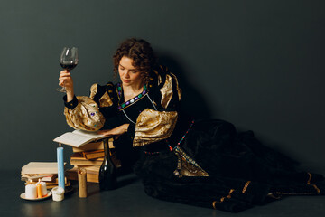 Fototapeta premium Young adult woman dressed in a medieval dress sitting near ancient books and candles with bottle of red wine and holding a glass of wine in dark