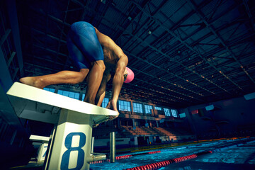 Ready to dive. Low angle view of young muscular man, swimmer, wearing goggles and cap, in low position on starting block in swimming pool. Concept of aquatic sport, preparation to competition, energy.