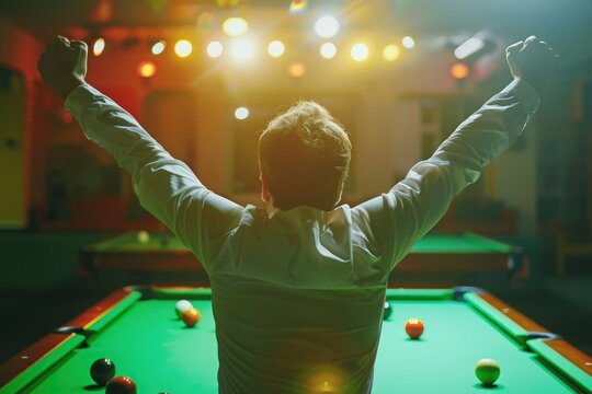 A pool player stands with arms raised in victory, illuminated by spotlights, after a successful game.