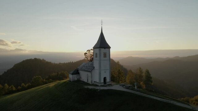 Aerial view of St. Primoz church at sunrise on a hill with mountain peaks in Julian Alps, Jamnik, Slovenia.