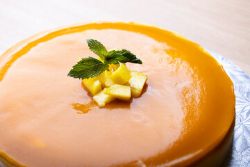 A beautifully glazed mango cheesecake topped with a dollop of mango puree and fresh mint leaves. The cake is presented on a silver cake board, capturing its smooth and shiny surface. 