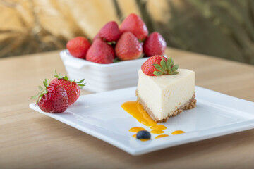 A slice of creamy cheesecake topped with a fresh strawberry, served on a white plate with a drizzle of fruit sauce. A bowl of fresh strawberries is in the background, adding vibrant color to the dish