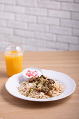 Delicious plate of biryani rice with tender chicken pieces, served with a side of yogurt garnished with pomegranate seeds, and accompanied by a refreshing orange juice in a plastic cup.