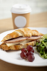 A freshly baked croissant sandwich filled with ham and cheese, served on a white plate with a side of mixed green salad and a few red grapes. A light and appetizing meal perfect for brunch or lunch.