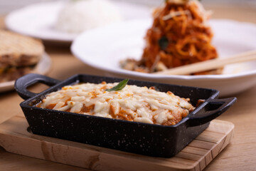 A delicious, cheesy lasagna served in a black ceramic dish, topped with melted mozzarella and garnished with a basil leaf. The dish rests on a wooden board with salt and pepper shakers and cutlery
