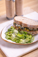 A wholesome sandwich filled with spinach, corn, and cheese, served on whole grain bread with a side of fresh green salad. The sandwich is placed on a rustic white plate with salt and pepper shakers