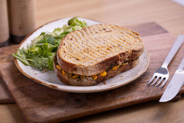 A wholesome sandwich filled with spinach, corn, and cheese, served on whole grain bread with a side of fresh green salad. The sandwich is placed on a rustic white plate with salt and pepper shakers