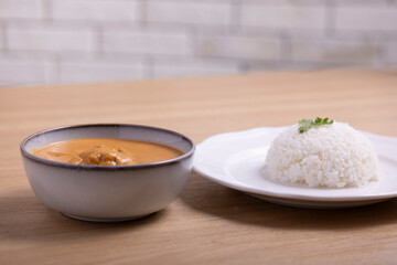 Delicious bowl of creamy butter chicken served with a mound of steamed white rice garnished with cilantro on a white plate, accompanied by a refreshing orange juice in a plastic cup. 