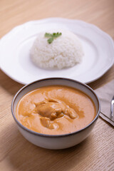 Delicious bowl of creamy butter chicken served with a mound of steamed white rice garnished with cilantro on a white plate, accompanied by a refreshing orange juice in a plastic cup. 