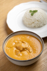Delicious bowl of creamy butter chicken served with a mound of steamed white rice garnished with cilantro on a white plate, accompanied by a refreshing orange juice in a plastic cup. 