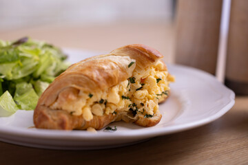 A breakfast spread featuring a croissant stuffed with scrambled eggs and herbs, served with a side salad on a white plate. Accompanied by a takeaway coffee cup from Postcard café,set on a wooden table