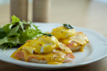 A delicious plate of eggs Benedict topped with rich hollandaise sauce, accompanied by a fresh side salad. The dish is beautifully presented on a white plate, placed on a vibrant outdoor table setting.