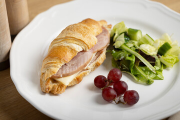 A freshly baked croissant sandwich filled with ham and cheese, served on a white plate with a side of mixed green salad and a few red grapes. A light and appetizing meal perfect for brunch or lunch.
