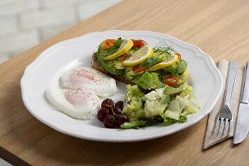 A delicious breakfast plate featuring poached eggs, avocado toast with lemon and tomato, a fresh salad, and a small bunch of grapes. Perfect for healthy eating and gourmet food themes.