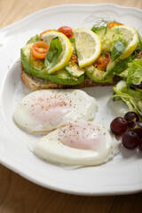 A delicious breakfast plate featuring poached eggs, avocado toast with lemon and tomato, a fresh salad, and a small bunch of grapes. Perfect for healthy eating and gourmet food themes.