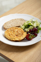 A delicious breakfast plate featuring a golden-brown omelette, two slices of whole grain bread, a fresh mixed green salad, red grapes, and a ripe strawberry. A nutritious and balanced meal ideal.