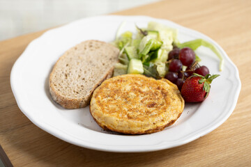 A delicious breakfast plate featuring a golden-brown omelette, two slices of whole grain bread, a fresh mixed green salad, red grapes, and a ripe strawberry. A nutritious and balanced meal ideal.