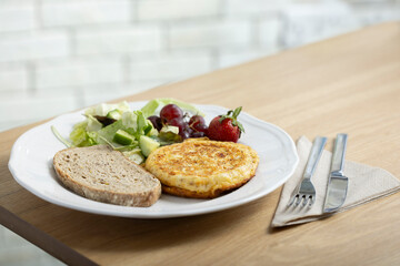 A delicious breakfast plate featuring a golden-brown omelette, two slices of whole grain bread, a fresh mixed green salad, red grapes, and a ripe strawberry. A nutritious and balanced meal ideal.