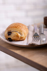 A mouthwatering close-up of a chocolate croissant, showcasing its golden, flaky layers and rich chocolate filling. Presented on a rustic white plate with a fork and knife on a napkin