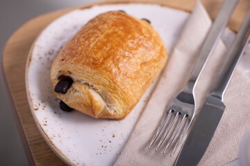A mouthwatering close-up of a chocolate croissant, showcasing its golden, flaky layers and rich chocolate filling. Presented on a rustic white plate with a fork and knife on a napkin