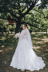 Wedding photography, portrait of a beautiful young happy smiling bride in a white long dress with a bouquet of flowers in a park outdoors in nature in greenery.