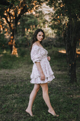 Beautiful young smiling happy barefoot brunette woman model in a summer sundress standing in a park outdoors. Photography, portrait, lifestyle.