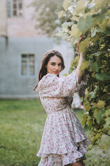 A beautiful young brunette woman model in a summer sundress stands near green grapes in the countryside outdoors. Close-up photography, portrait, lifestyle.