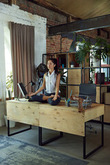 Woman, office worker sitting on desk surrounded by computer and office supplies, doing calming meditation in yoga lotus pose. Concept of office life, balance between work and sport, business