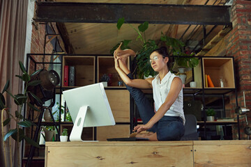Woman, employee in formal wear and glasses, sitting on desk and doing stretching exercises in front of computer. Physical well-being in modern work culture. Concept of balance between work and sport
