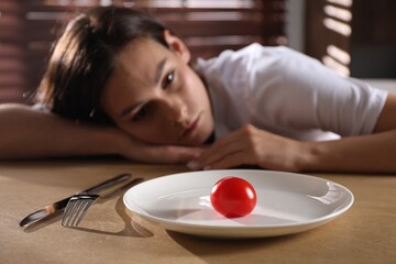 Eating disorder. Sad woman at wooden table with cutlery, tomato and plate indoors