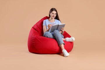 Beautiful young woman reading book on red bean bag chair against beige background