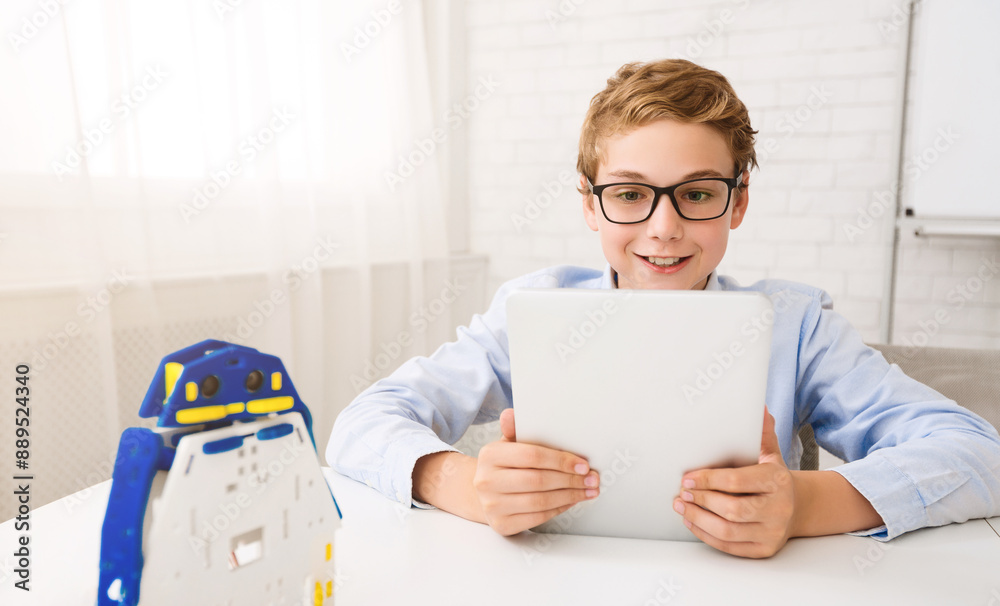 Poster A young boy with glasses sits at a desk and uses a tablet. He looks intently at the screen, smiling. There is a small robot toy on the desk in front of him.