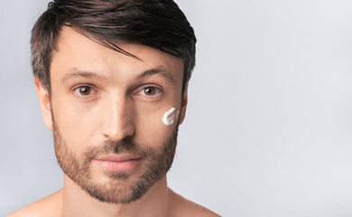 Obraz premium A man with a beard applies cream to his cheek while looking directly at the camera.