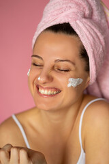 A young woman applying cream on her face looking happy, with wide smile and smooth skin and a woman standing in the bathroom doing her make up routine 