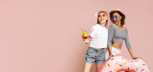 Two women in sunglasses, hats, and summery clothes pose together against a pink backdrop. One holds a cocktail and the other rests her hands on a large pink inflatable donut pool float.