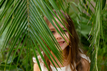 Fototapeta premium The face of a happy little girl surrounded by tropical leaves. Closeup portrait of a beautiful child with perfect skin and dark hair. Natural cosmetics, health, cleanliness, skin care, beauty concept
