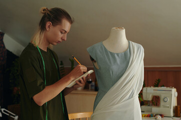 Young fashion designer standing near the mannequin and writing measurements in notebook