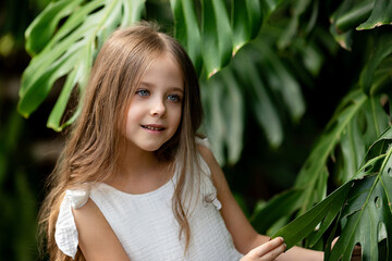 Fototapeta premium The little girl surrounded by tropical monstera leaves. Closeup portrait of a beautiful child with perfect skin and dark hair. Natural cosmetics, health, cleanliness, skin care, beauty concept