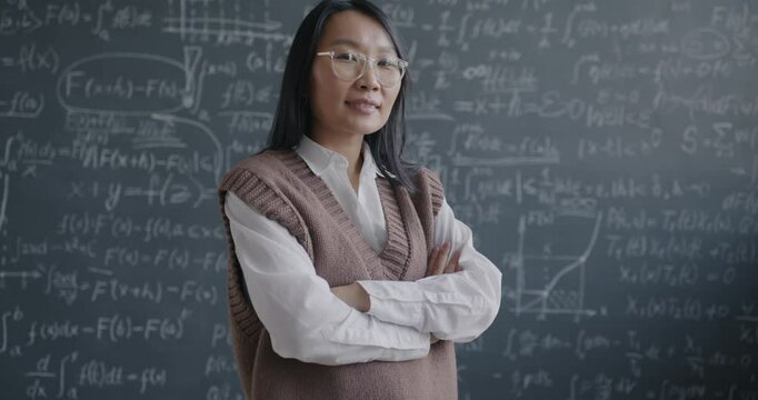 Zoom-in portrait of young Asian lady student standing in classroom in front of chalkboard with formula smiling looking at camera. Education and people concept.