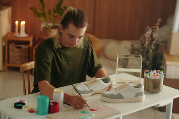 Designer sitting at table and decorating sneakers with paints in fashion style