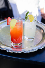 two highball glasses, one with lemonade and one with strawberry lemonade, being served on silver tray at outdoor event